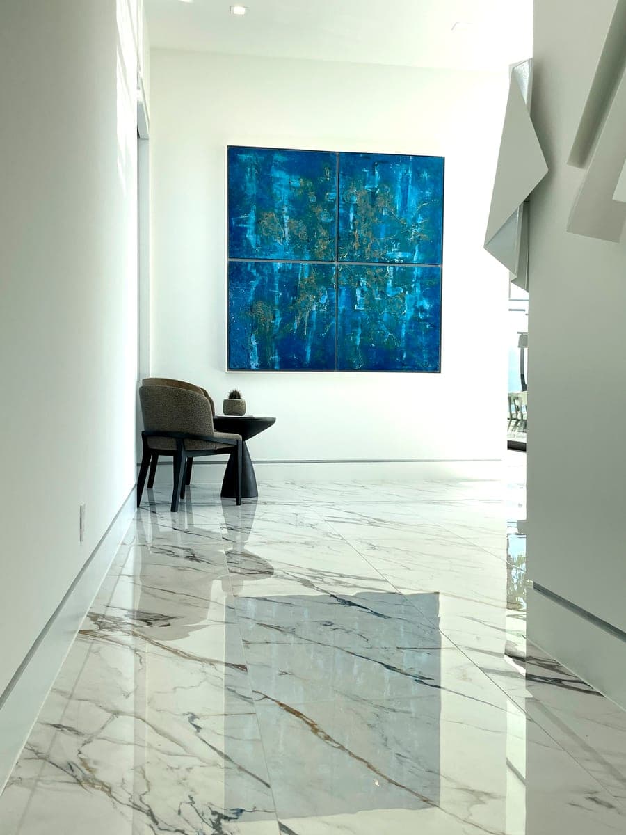 White hallway with a polished marble floor; a four-panel blue and gold work hangs above a grey armchair and a small black side table.